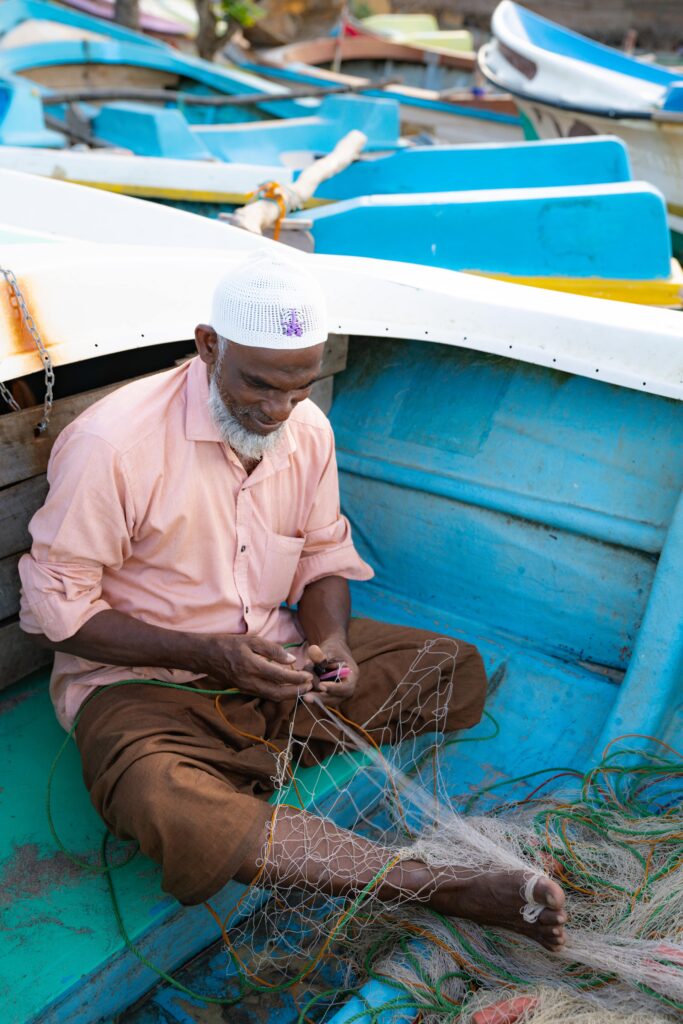 An elderly man wearing a white cap and pink shirt sits in a blue boat, mending a fishing net with his hands—a glimpse of local life among the colorful boats, and one of the authentic things to do in Arugam Bay.