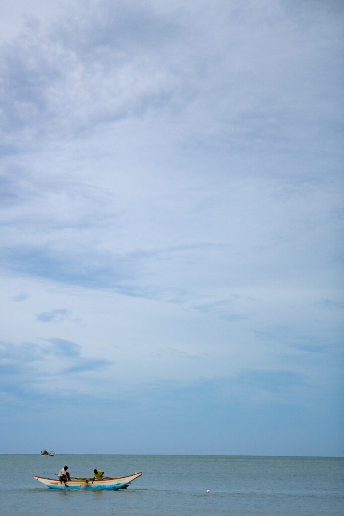 A small boat with two people floats on calm blue water under a wide, cloudy sky—capturing the serene beauty often found among Things To Do In Arugam Bay—with another boat faintly visible in the distance.