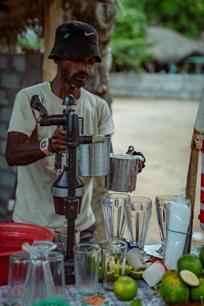 A man wearing a black hat and light t-shirt squeezes juice with a metal press at an outdoor stand—one of the vibrant things to do in Arugam Bay. Fresh limes, blenders, glasses, and a red container are on the table in front of him.