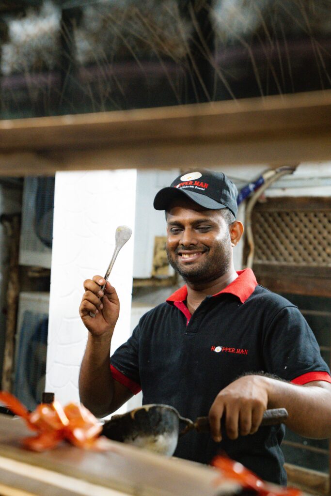 A smiling man wearing a black cap and shirt stirs food with a ladle in one hand and holds a spoon in the other, standing behind a counter in what looks like a kitchen or food stall—a must-see for those seeking local Things To Do In Arugam Bay.
