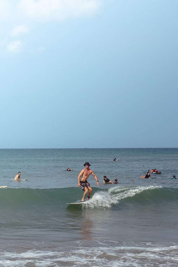 A person surfing a small wave near the shore while several others swim and surf in the ocean under a blue sky—one of the top things to do in Arugam Bay.