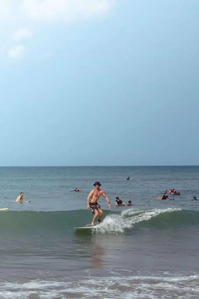 A person surfing a small wave near the shore while several others swim and surf in the ocean under a blue sky—one of the top things to do in Arugam Bay.
