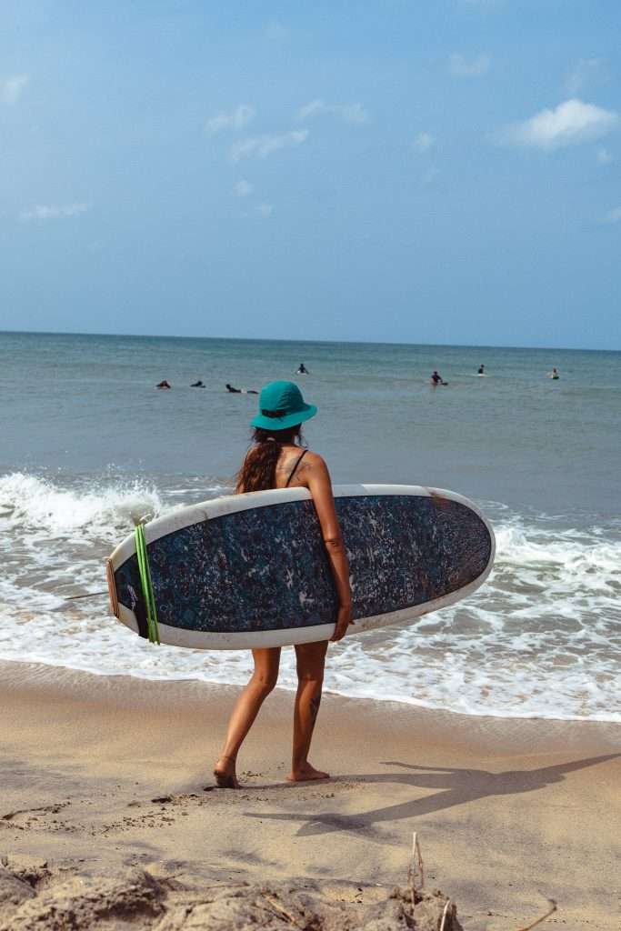 A woman in a green hat stands on the beach with her surfboard, gazing at the ocean where others enjoy the waves under a partly cloudy sky—one of the top things to do in Arugam Bay.