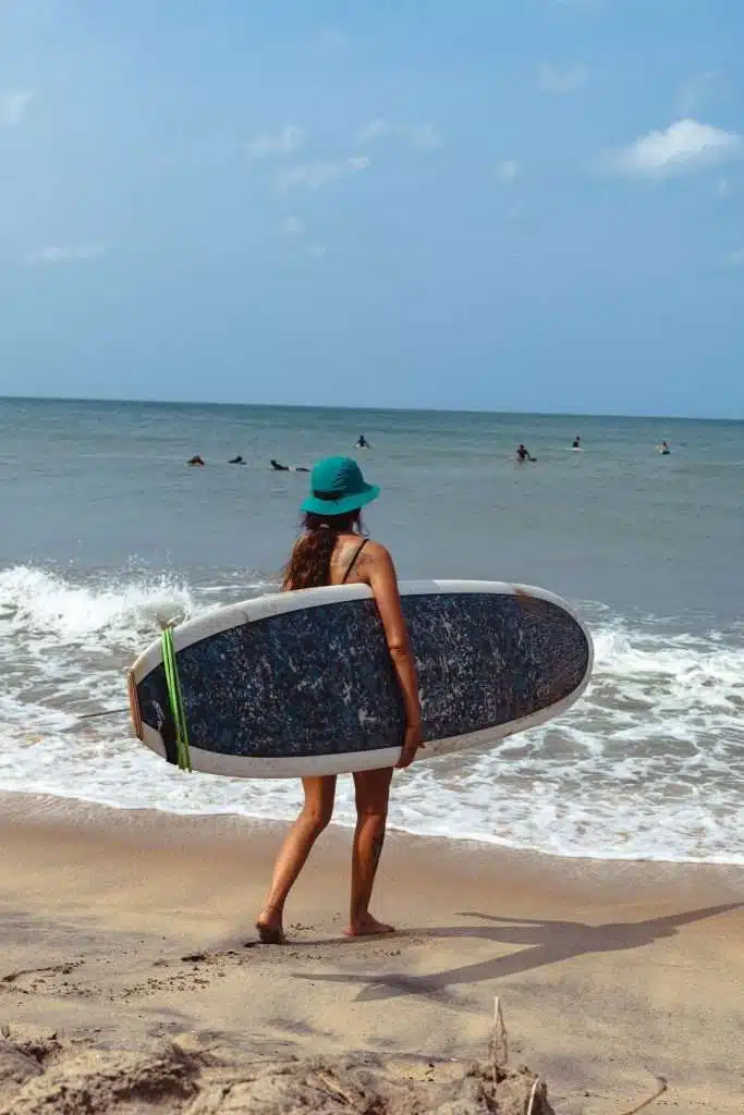 A woman in a green hat stands on the beach with her surfboard, gazing at the ocean where others enjoy the waves under a partly cloudy sky—one of the top things to do in Arugam Bay.