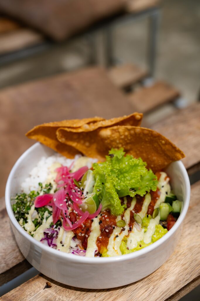 A white bowl filled with rice, fresh greens, pickled onions, diced vegetables, sesame seeds, and creamy sauce topped with crispy wonton chips sits on a wooden table—a vibrant meal to enjoy after exploring Things To Do In Arugam Bay.