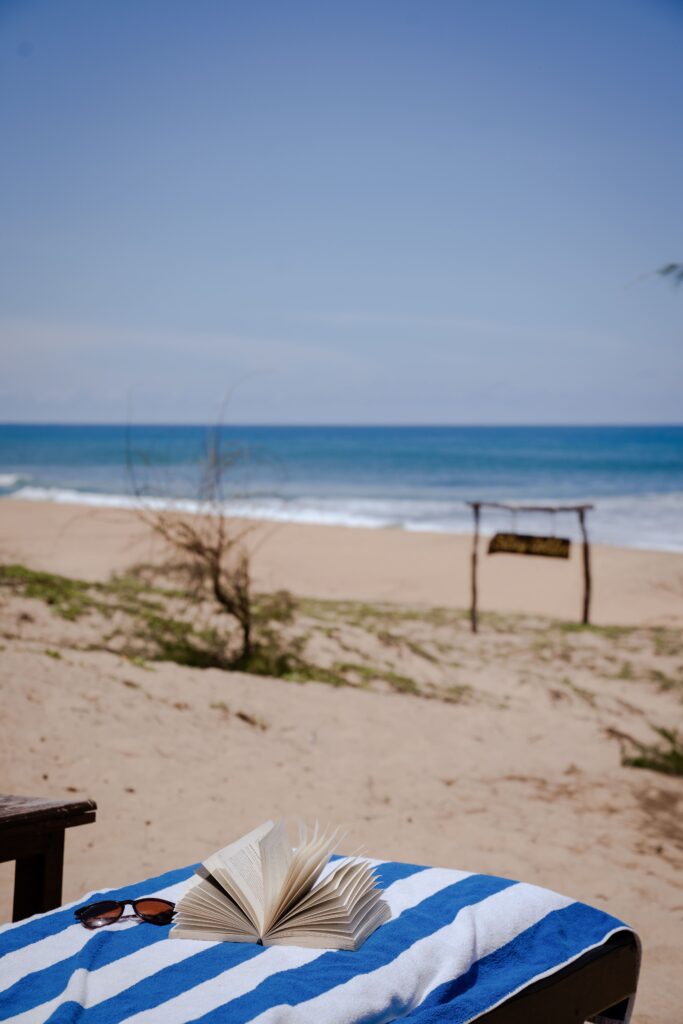 A book and sunglasses rest on a blue-and-white striped towel on a sandy beach, with the ocean and a wooden sign in the background—a perfect scene for those seeking Things To Do In Arugam Bay under a clear blue sky.