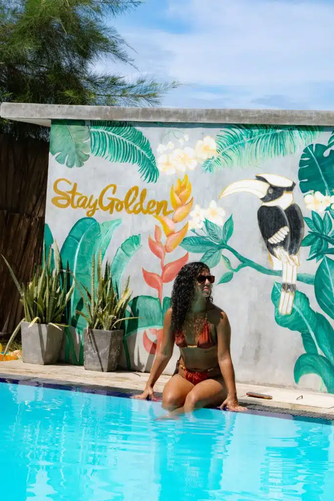 A woman in a brown bikini sits by a pool, her legs in the water. Behind her, a vibrant mural with flowers and "Stay Golden" captures the sunny vibe—perfect for your list of things to do in Arugam Bay. Potted plants add tropical charm.