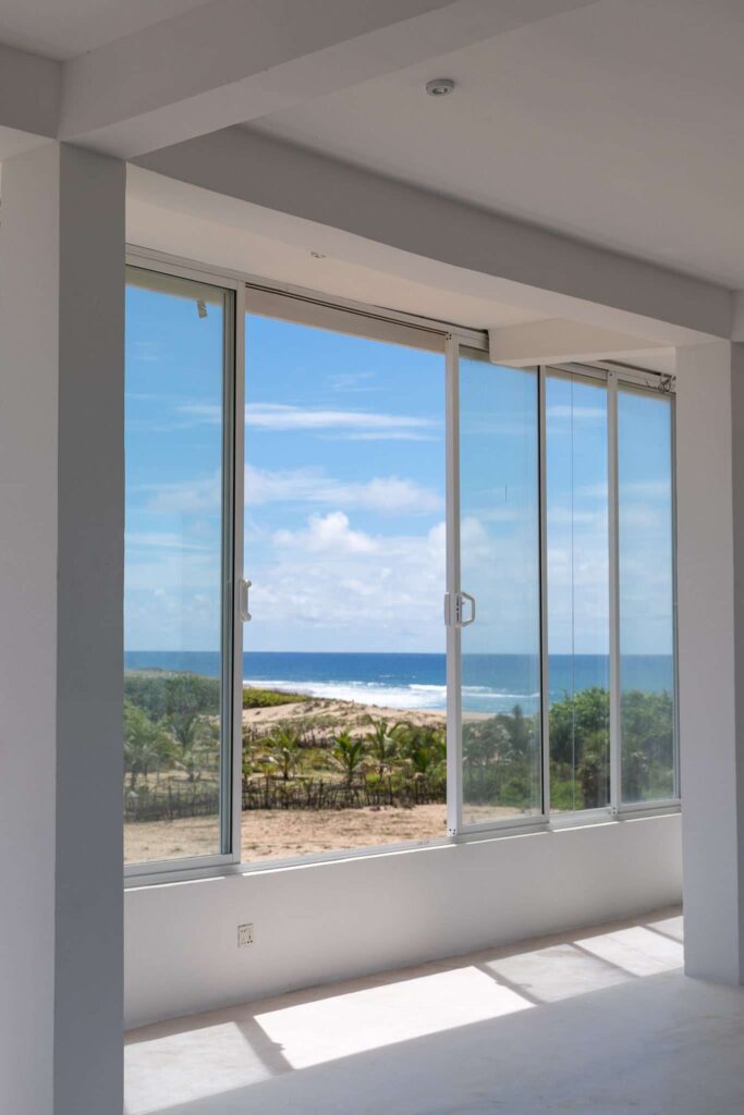 Large floor-to-ceiling glass windows in a white room offer a clear view of a sandy beach, palm trees, and the blue ocean—ideal for relaxing after exploring the many Things To Do In Arugam Bay under a partly cloudy sky.