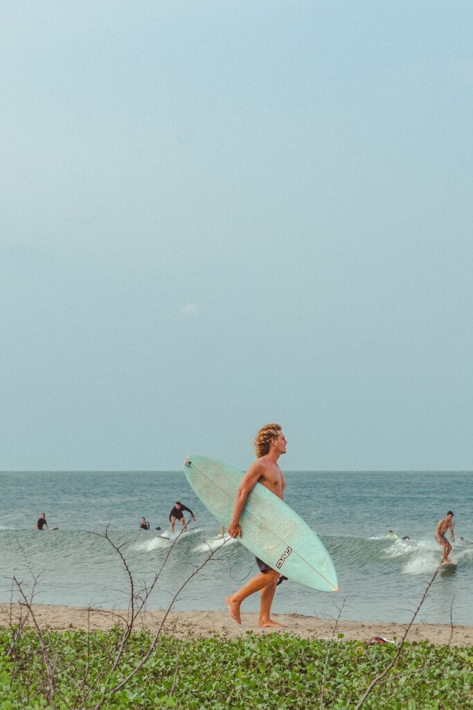 A person with curly blond hair carries a surfboard along a sandy beach—one of the classic things to do in Arugam Bay. In the background, several people are surfing under a clear sky, with green vegetation in the foreground.