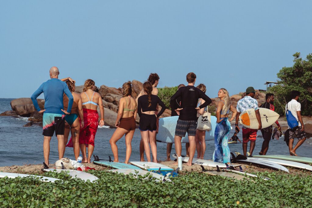 A group of people with surfboards stand on a grassy shore, facing the ocean and rocks—one of the top things to do in Arugam Bay. Some wear swimsuits or rash guards, looking toward the water, preparing to surf under a clear sky.