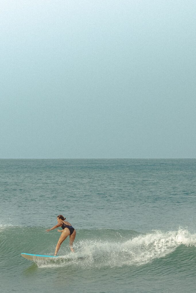 A person in a dark swimsuit rides a small wave on a surfboard in the ocean under a clear sky, enjoying one of the top things to do in Arugam Bay.