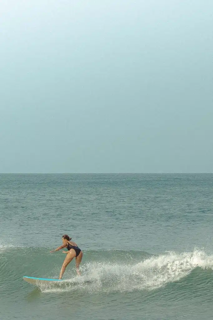 A person in a dark swimsuit rides a small wave on a surfboard in the ocean under a clear sky, enjoying one of the top things to do in Arugam Bay.