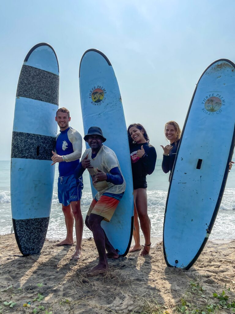 Four people holding up blue surfboards stand on a sandy beach, smiling and posing with the ocean behind them—a sunny, cheerful scene that highlights one of the top Things To Do In Arugam Bay.