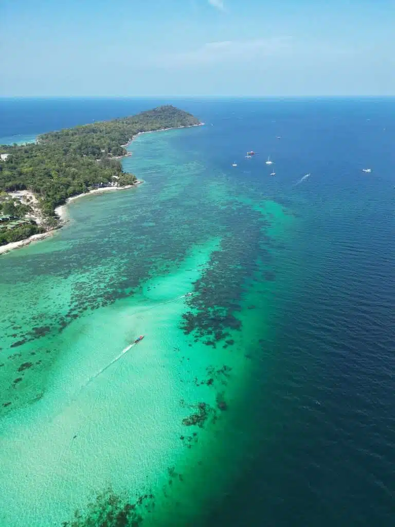 Aerial view of the Koh Lipe coastline with lush green forest meeting turquoise waters, hinting at things to do in Koh Lipe. Boats are scattered across the sea, with an island in the background under a clear blue sky.