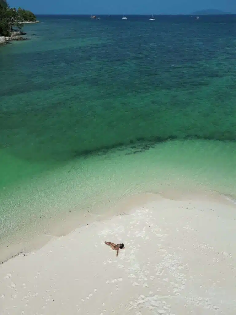 A person lies on a pristine white sandy Sunrise beach in Koh Lipe, with turquoise waves gently reaching the shore. The sea extends to the horizon, dotted with distant sailboats under a clear blue sky. 