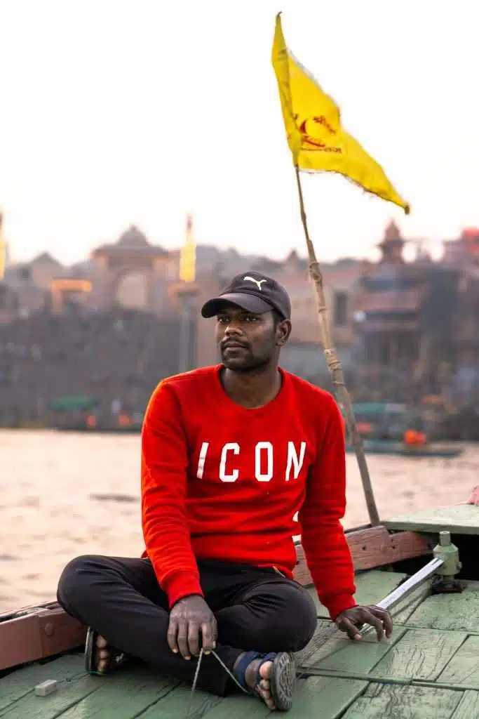 A man in a red "ICON" sweatshirt and black cap sits cross-legged on a wooden boat by the river, with a yellow flag and a blurred cityscape behind him—an idyllic moment from a Varanasi 2-Day Itinerary.