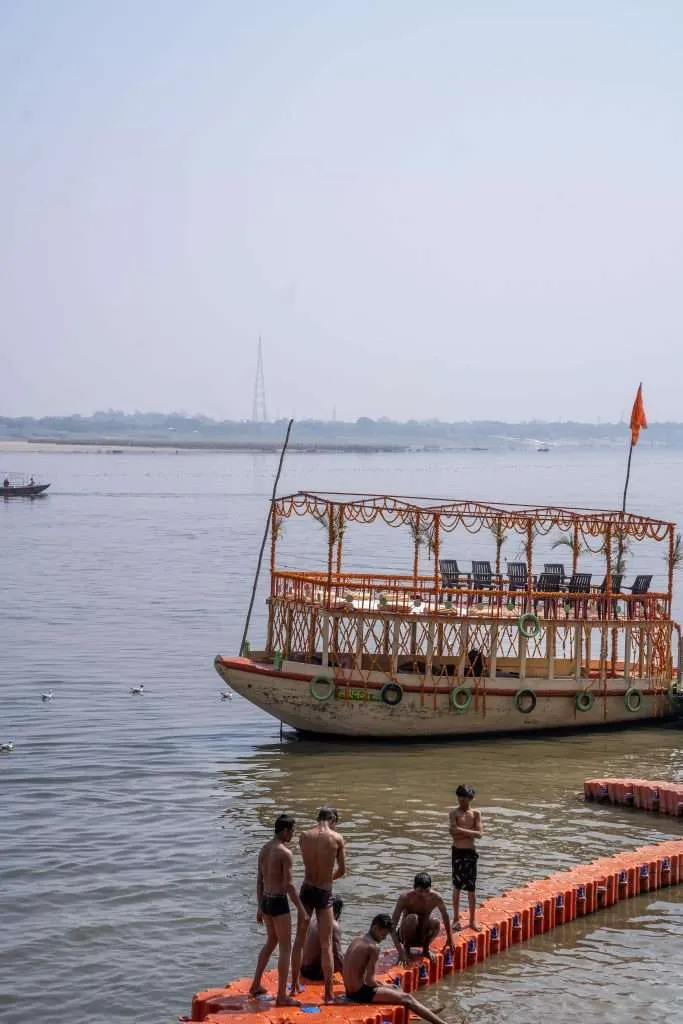 A decorated boat with orange garlands floats on a river, while a group of boys in swimwear gather on an orange floating platform near the water’s edge—an ideal scene to include in your Varanasi 2-Day Itinerary under a clear, blue sky.