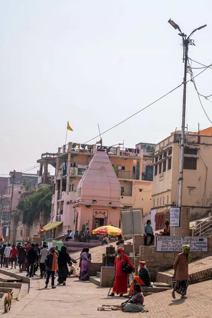 A busy street scene in India, perfect for a Varanasi 2-Day Itinerary, with people walking and sitting near a small pink temple, surrounded by buildings, street signs, and a streetlight on a hazy day. Vendors and a dog are also visible.