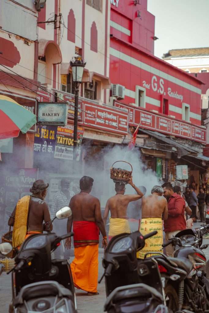 A group of men, some in traditional attire, walk down a street market in Varanasi. One holds a large basket on his head as a haze of smoke surrounds them. In the background, shops and the hotel "G.S. Residency" are visible with motorcycles parked along the street. Is Varanasi worth visiting? Absolutely.