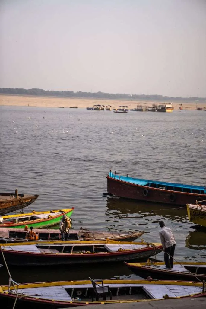 Several colorful wooden boats are docked along the edge of a wide river in Varanasi, with people on a few boats. The opposite riverbank and hazy sky create a serene backdrop, perfect for your Varanasi 2-Day Itinerary.