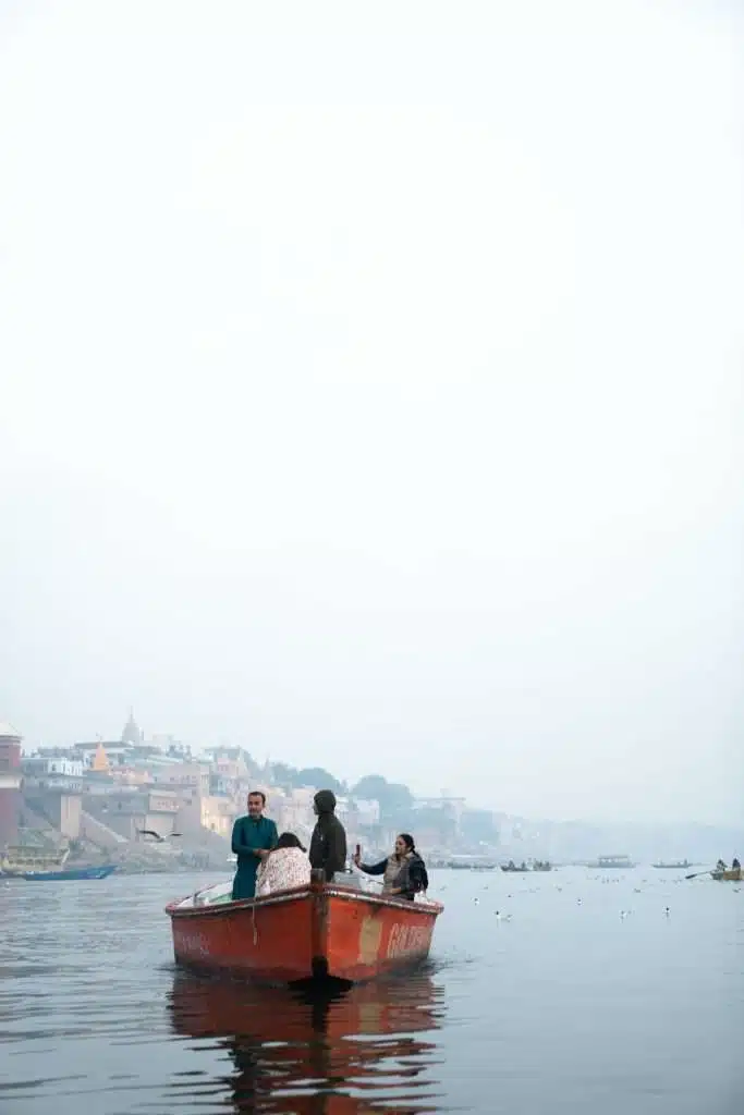 A small red boat with four people aboard floats on a calm river, reminiscent of the tranquil scenes that make one ponder: Is Varanasi worth visiting? A foggy cityscape with colorful buildings and distant boats is visible in the background, under a hazy sky.