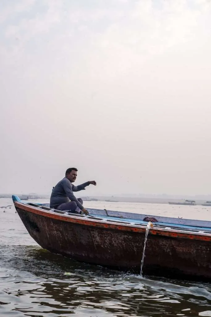 A man sits on the edge of a wooden boat floating on a river, scooping water with his hand—a tranquil scene that could be part of any Varanasi 2-Day Itinerary. The hazy sky and distant boats add to the calm atmosphere.