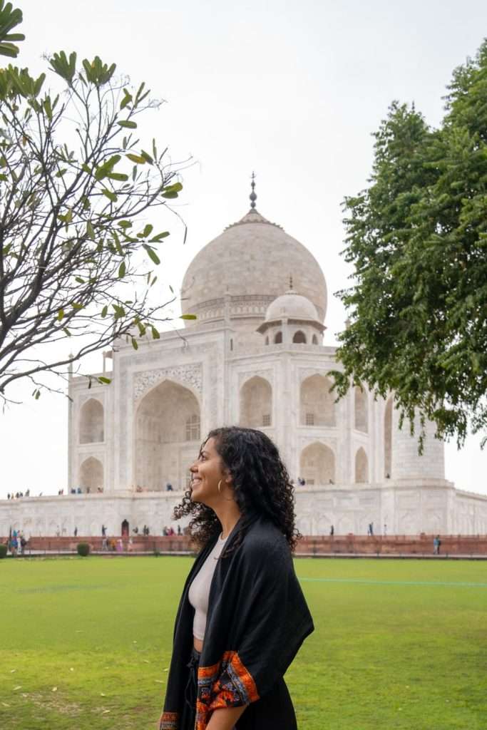 A person with curly hair, wearing a dark shawl with orange patterns, stands smiling to the side. The majestic white marble Taj Mahal stands prominently in the background, framed by green trees and an expansive lawn. 