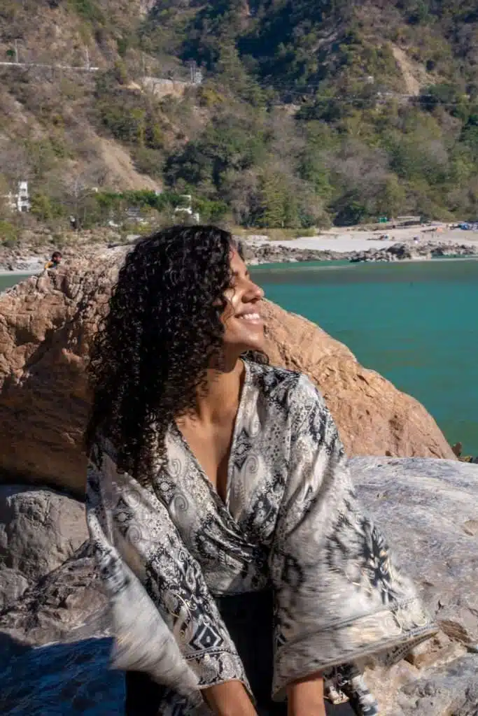 A person with curly hair and a patterned shawl smiles while sitting on a rock in front of a picturesque landscape featuring green hills, blue water, and a sandy beach in Rishikesh India