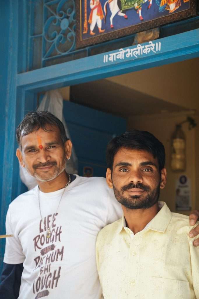 Two men standing together in front of a blue doorway. The man on the left is wearing a white T-shirt with text and has a red mark on his forehead. The man on the right is wearing a beige shirt. Both men are smiling and looking at the camera, perhaps discussing if India is safe for solo female travelers.