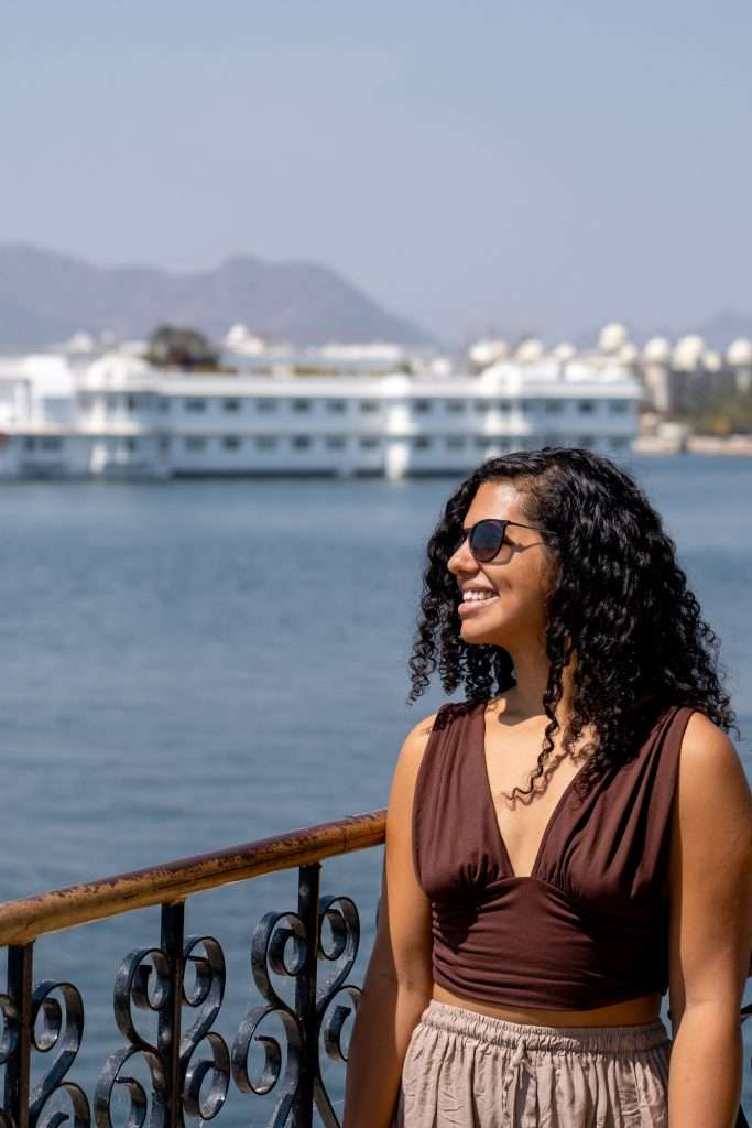 A woman with curly hair and wearing sunglasses and a sleeveless top stands on a balcony with ornate railings. She is smiling and looking off to the side. In the background, a large boat and mountains can be seen on a calm body of water under a clear sky - Udaipur India