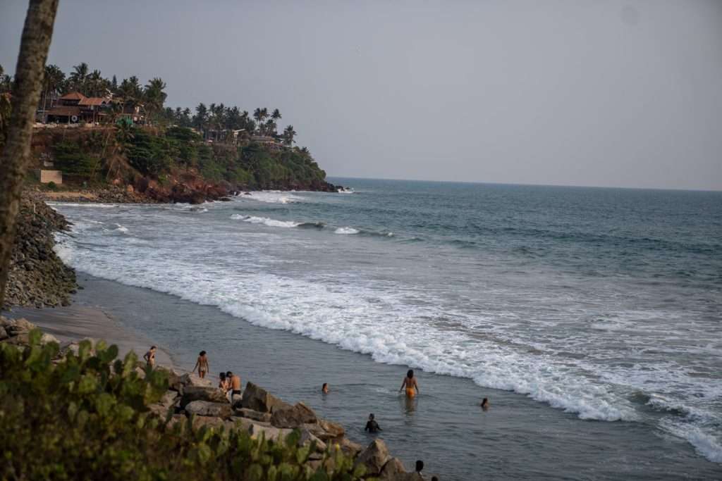 A sandy beach in Varkala India with gentle waves where several people are swimming and standing in the shallow water. In the background, there are rocky cliffs with palm trees and some buildings nestled among them.