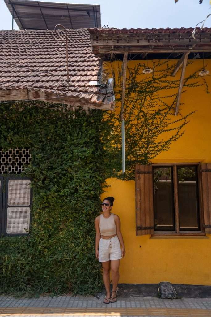 A person in Fort Kochi India, wearing sunglasses, a beige tank top, and white shorts stands against a vibrant building, pondering the question: Is India safe for solo female travellers? The building has a wall covered in green ivy on the left and a yellow wall with a window on the right. The roof is made of red tiles.