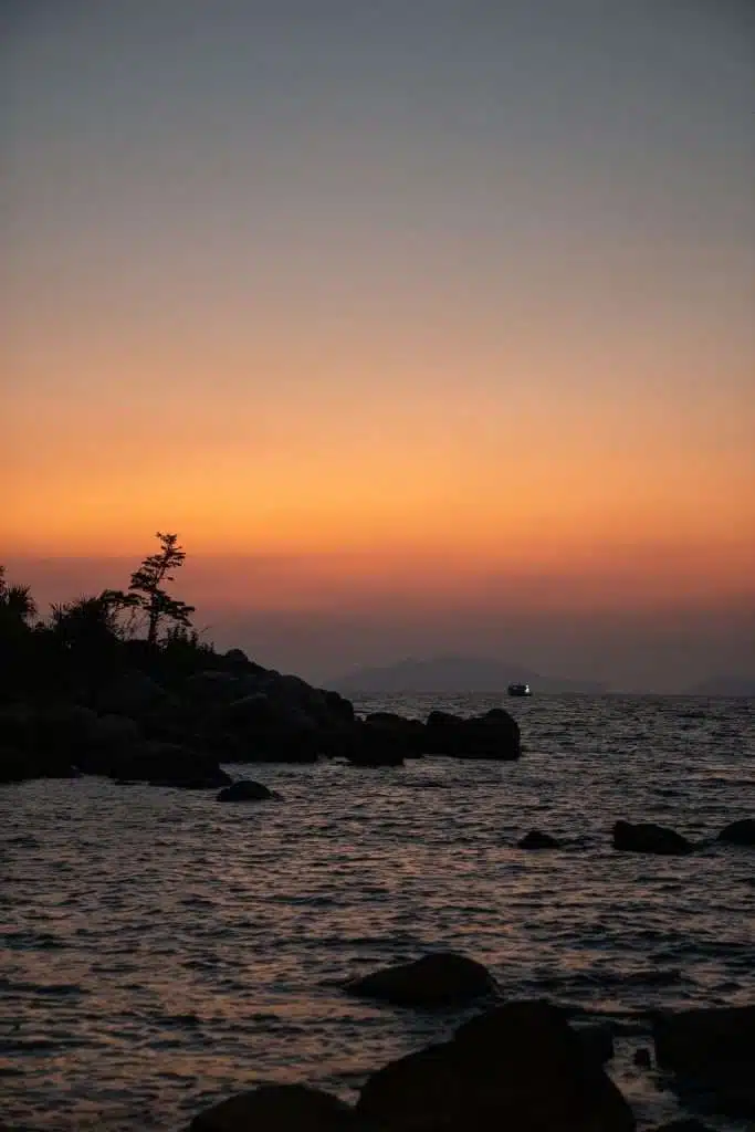 A tranquil sunset over a rocky shoreline in Sunset in Koh Lipe, with the sky painted in gradients of orange and blue. A distant boat with lights over the calm water is silhouetted against the horizon, while a tree leans gracefully over the rocks on the left