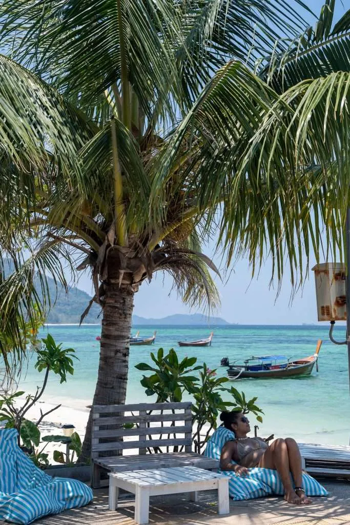 A person relaxes on a striped beanbag under a palm tree on a sandy beach in Koh Lipe. Crystal-clear turquoise water and boats are in the background, with distant green hills under a clear blue sky.