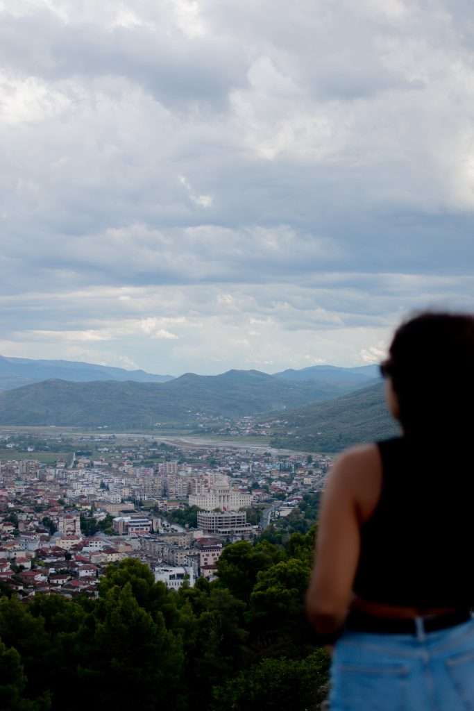 A person with long hair, wearing a sleeveless top and jeans, stands looking out over a cityscape nestled among mountains. The overcast sky is scattered with clouds as the city below showcases a mix of buildings and greenery.