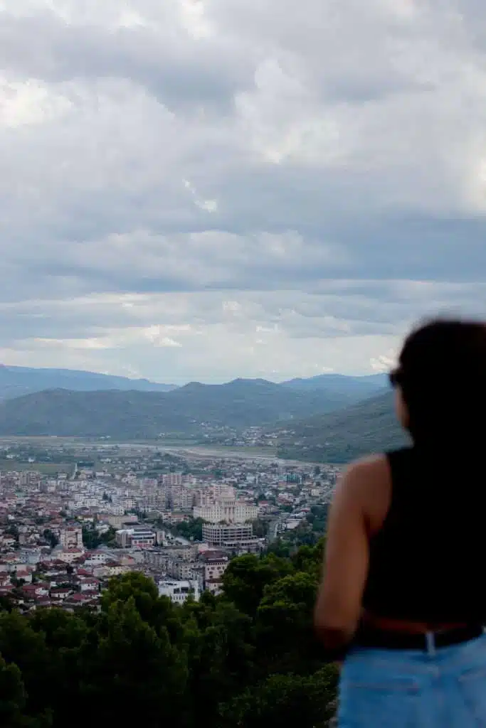 A person with long hair, wearing a sleeveless top and jeans, stands looking out over a cityscape nestled among mountains. The overcast sky is scattered with clouds as the city below showcases a mix of buildings and greenery.