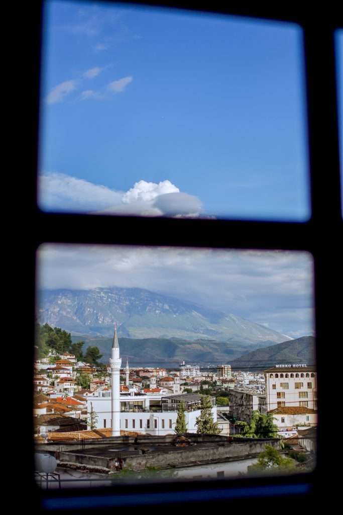 View through a window shows a picturesque town with a prominent white minaret, set against a backdrop of mountains and a blue sky with clouds. This serene scene is an inviting part of any 1-Week Albania Itinerary, showcasing various buildings and lush greenery.