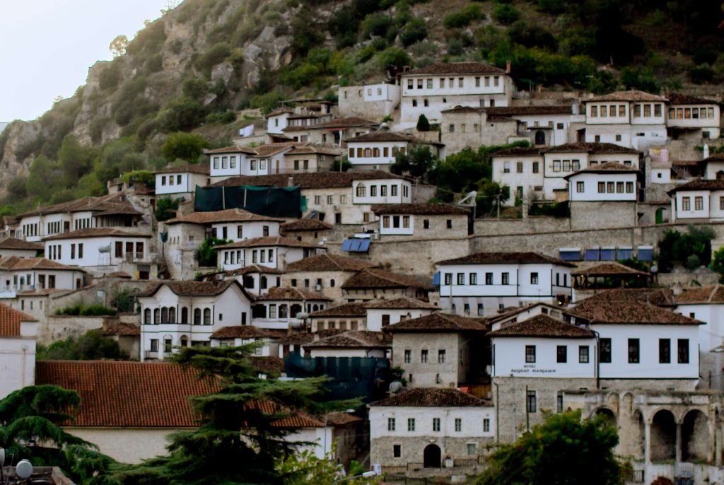 A hillside town features white houses with brown roofs clustered together. A fortress with a flag sits atop the hill surrounded by lush green trees. The sky above is overcast, adding to the serene ambiance of the quaint village, a perfect stop on your 1-Week Albania Itinerary.