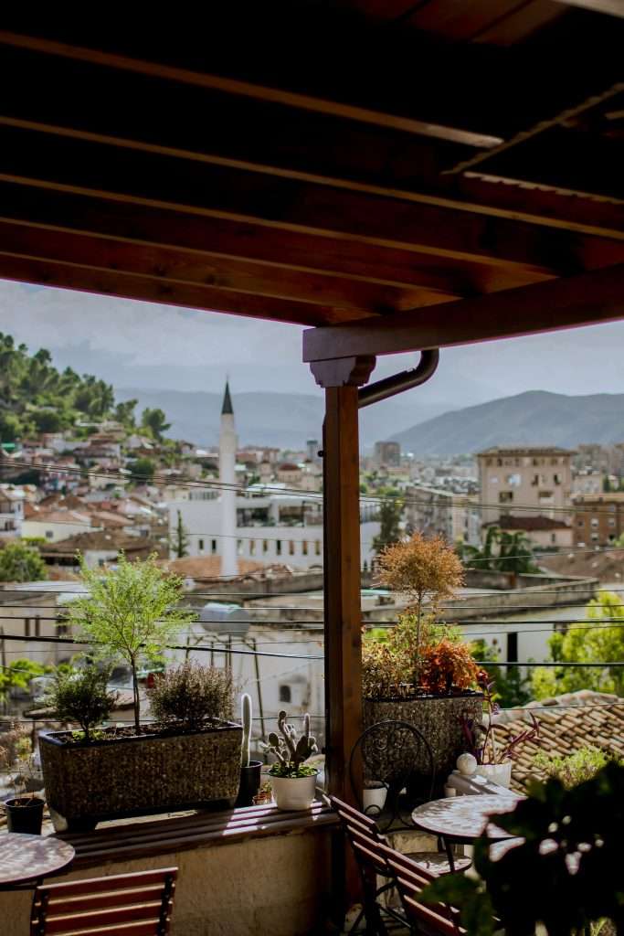 A cozy terrace with potted plants, perfect for unwinding during your 1-Week Albania Itinerary, overlooks a scenic town with scattered buildings, including a minaret. The background features rolling hills and mountains under a partly cloudy sky.