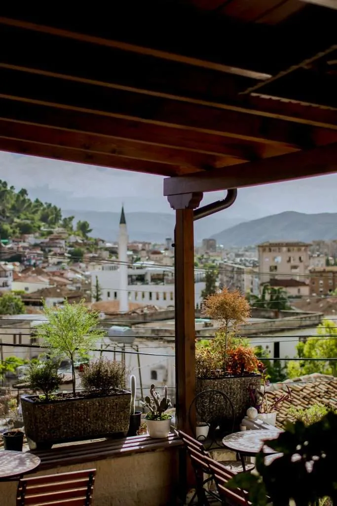 A cozy terrace with potted plants, perfect for unwinding during your 1-Week Albania Itinerary, overlooks a scenic town with scattered buildings, including a minaret. The background features rolling hills and mountains under a partly cloudy sky.