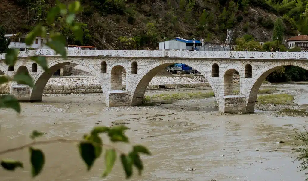 A picturesque stone bridge with arched supports spans a muddy river, set against a backdrop of green, tree-covered hills under a partly cloudy sky. Nearby, small buildings and construction equipment hint at new developments—a perfect sight to include in your 1-Week Albania Itinerary.