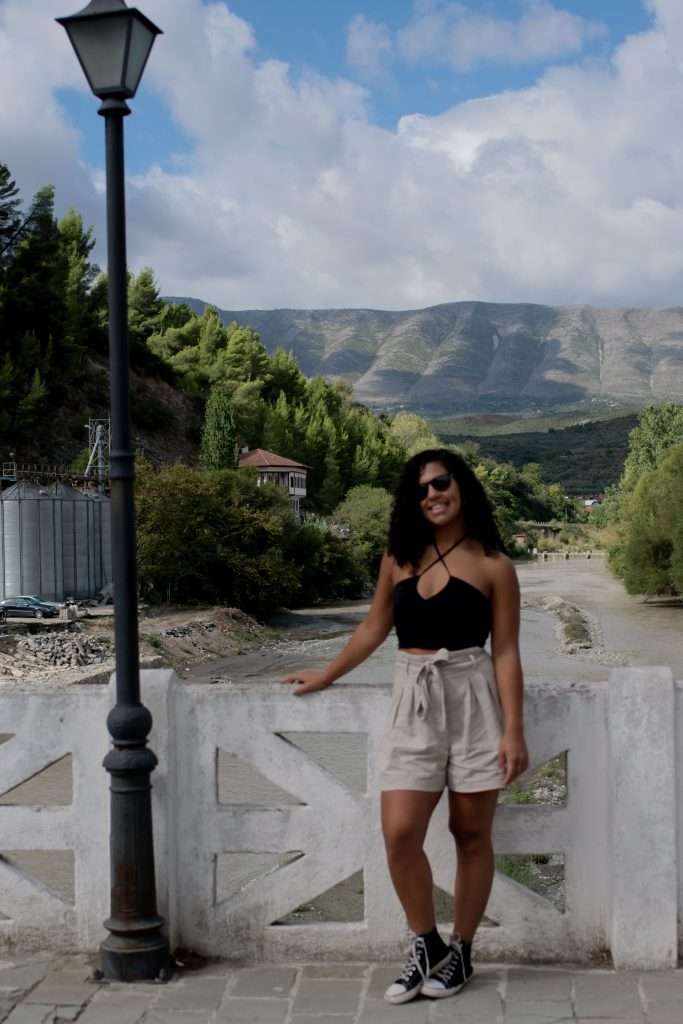 A woman with curly hair and sunglasses is standing on a bridge, leaning against a white railing. She is wearing a black top and beige shorts. The backdrop looks straight out of a 1-Week Albania Itinerary, featuring scenic mountains, lush greenery, and a cloudy sky in Berat Albania.