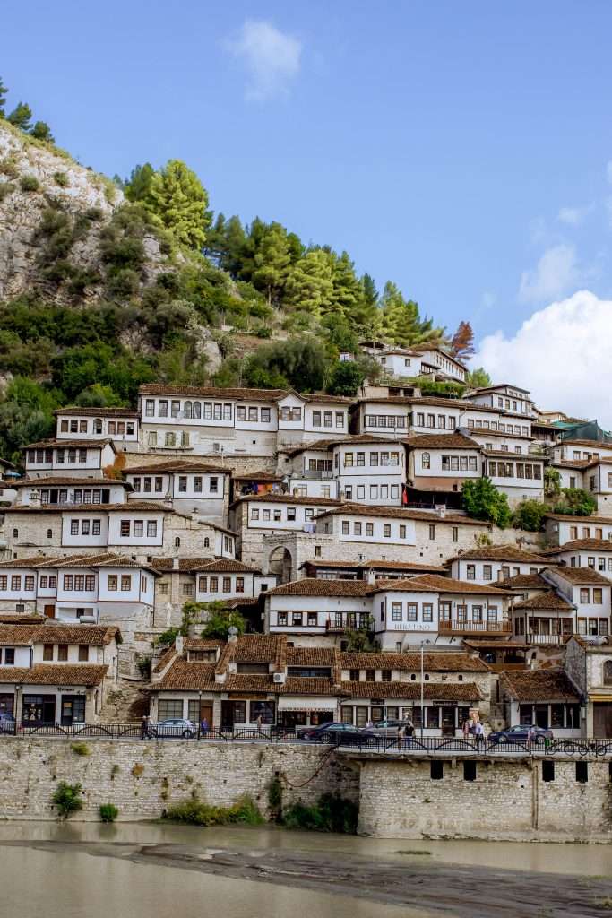 A photo showcasing traditional white Ottoman-style houses with wooden shutters built on the hillside of Berat, Albania—an essential stop on a 1-week Albania itinerary. The houses rise in tiers, surrounded by lush greenery under a bright blue sky with a few scattered clouds. The foreground features a river.