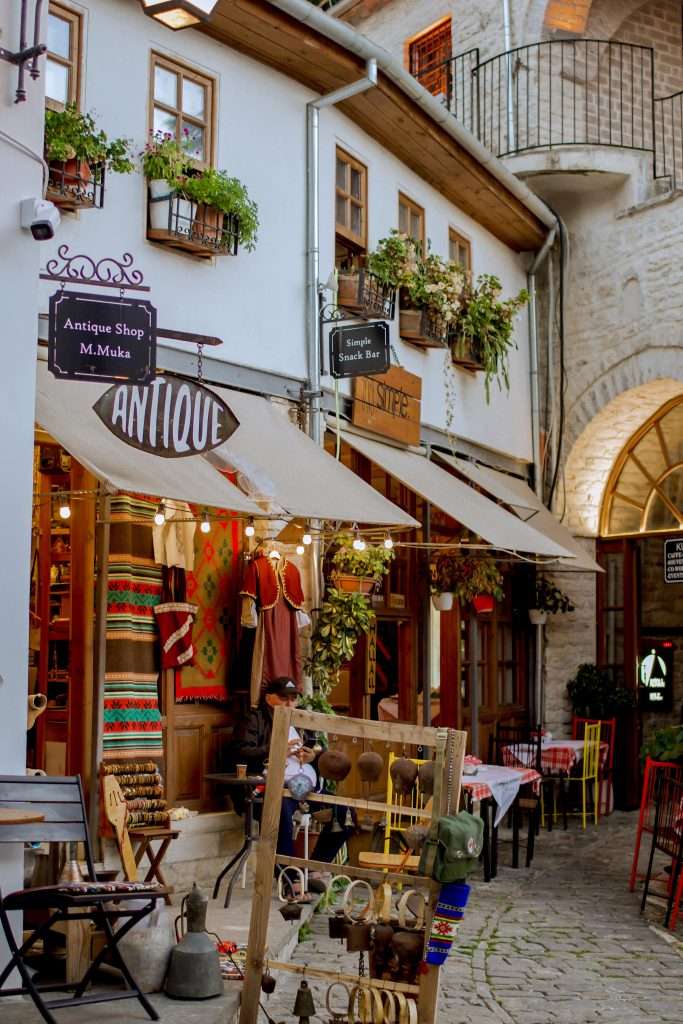 A charming street scene reminiscent of a week in Albania, featuring a cozy antique shop and cafe. Hanging plants and colorful textiles grace the shop front under a beige canopy. Cobblestone paths and wooden chairs enhance the quaint atmosphere, inviting exploration in Gjirokaster Old Town, Albania