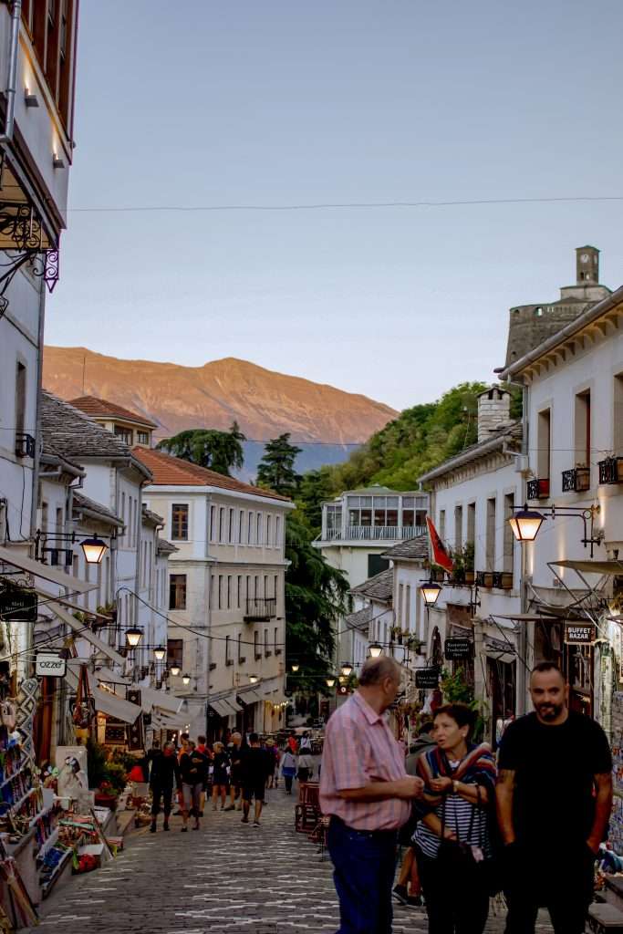 A bustling, narrow street in a charming, historic town with people walking and chatting. Stalls line the street with souvenirs and goods. Traditional white buildings adorned with lanterns and balconies frame the scene, part of your unforgettable 5-Day Albania Itinerary. Picturesque mountains under a clear sky loom in the background in Gjirokaster