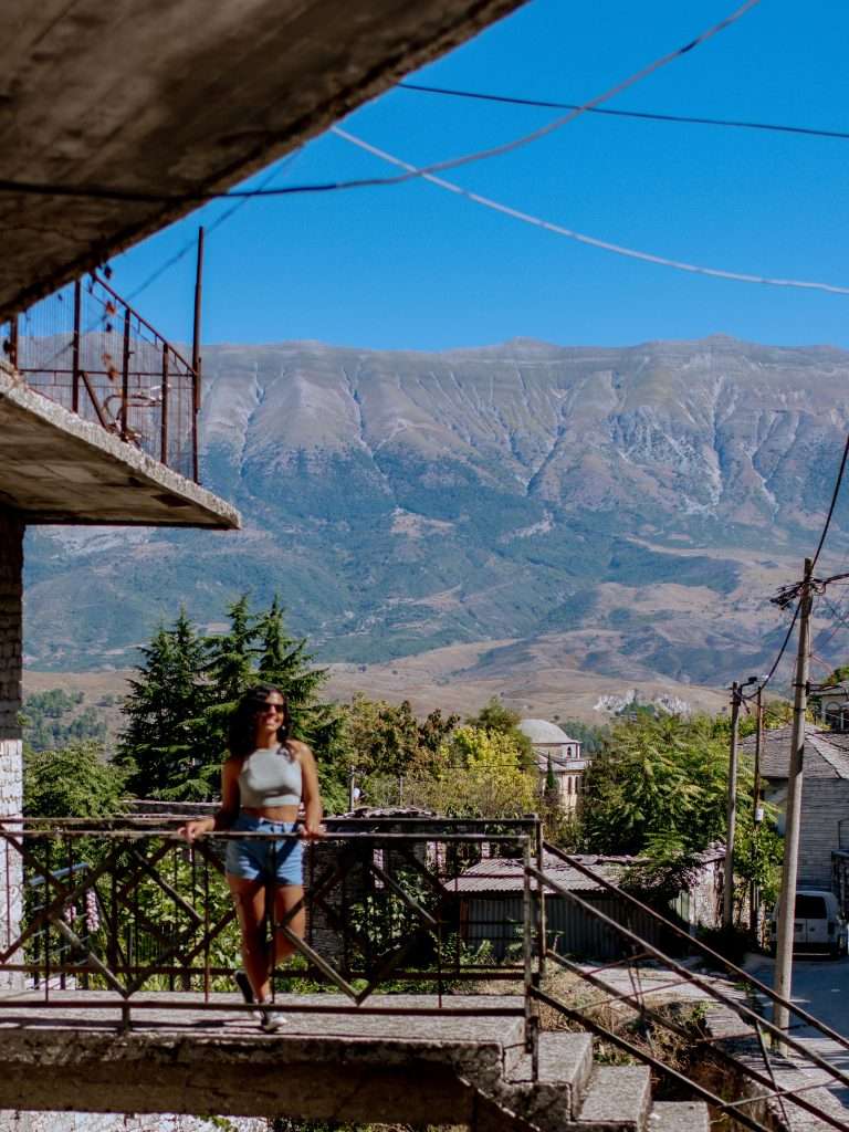 A person stands on the lower level of an old, concrete structure, posing near a metal railing. Behind them, a scenic mountainous landscape is visible with trees and buildings at the base. The clear blue sky in Gjirokaster enhances the picturesque setting, making it a perfect stop on a 1-Week Albania Itinerary.