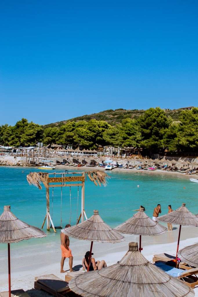 A beach scene with turquoise water, white sand, and several thatched sun umbrellas. A wooden sign reads "Poda E Ksamil". People are lounging, swimming, and walking around. Pine-covered hills and a crowded shoreline with sunbeds add charm to this picturesque stop on any 5-Day Albania Itinerary.