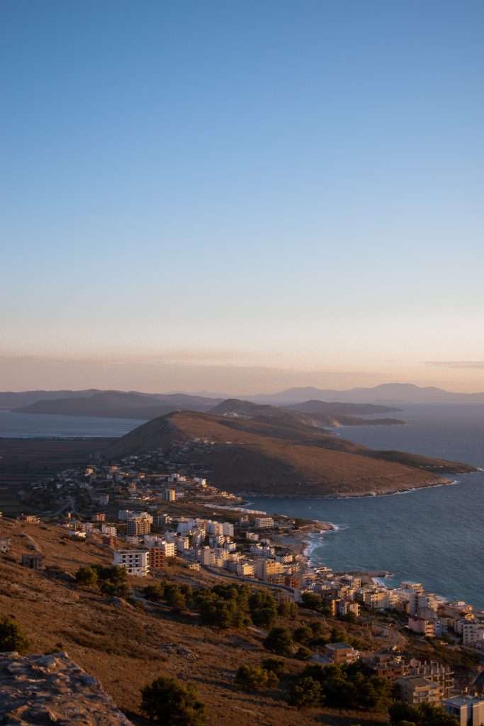 A scenic view of a coastal city at sunset at Lekuresi Castle in Saranda Albania with buildings and houses lining the shoreline. The city is bordered by hills and a calm sea, with distant mountains on the horizon under a clear, blue sky.