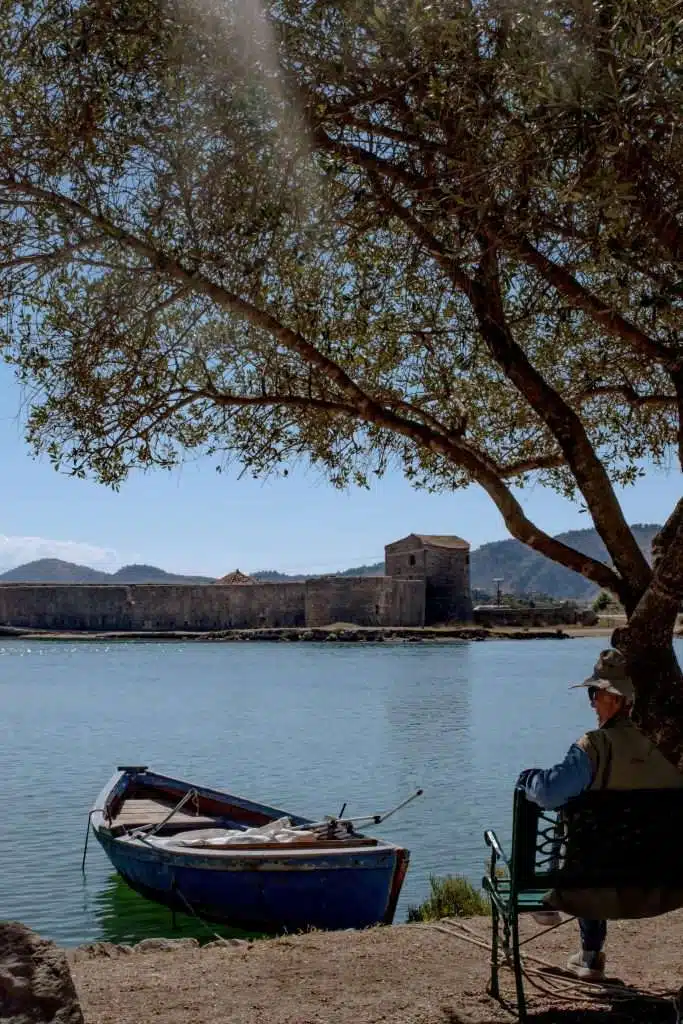 A peaceful lakeside scene with an elderly person sitting on a bench under a tree, gazing at a small boat anchored on calm waters. In the background, an old stone building is visible on the opposite shore, with hills and clear skies adding to the tranquility—a perfect moment in your 1-Week Albania Itinerary - Butrint National Park