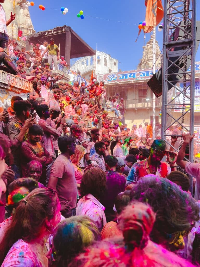 A large crowd celebrates Holi, covered in colorful powders, in a lively street scene near temples. People smile and cheer under vibrant decorations. Such festive scenes spark curiosity: Is India safe for solo female travellers?.